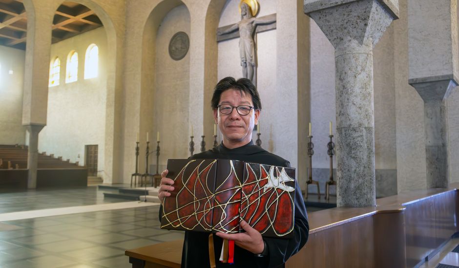 Mönch im schwarzen Gewand mit einem großen Buch in einer Kirche, im Hintergrund steht ein großes Kreuz
