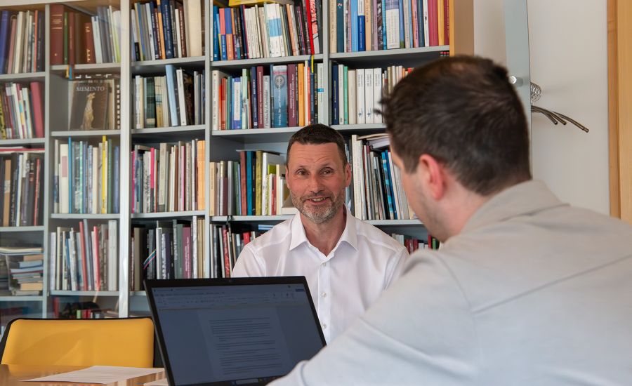 Zwei Männer unterhalten sich, im Hintergrund steht ein Buchregal, auf dem Tisch ist ein Laptop