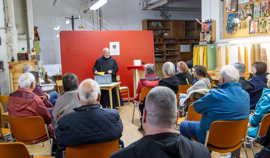 Mönch steht vor einer roten Rückwand und spricht vor einem Publikum