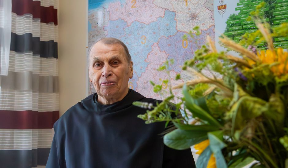 Älterer Mönch in einem Büro vor einer Landkarte, neben ihm ein Strauß Sonnenblumen